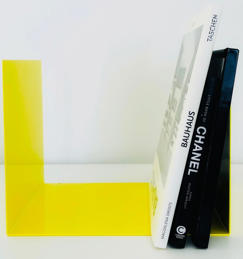 Eine Kollektion von Designbüchern, darunter Titel über das Bauhaus und Chanel, lehnt an einem leuchtend gelben Buchstützen aus Metall auf einem weißen Hintergrund